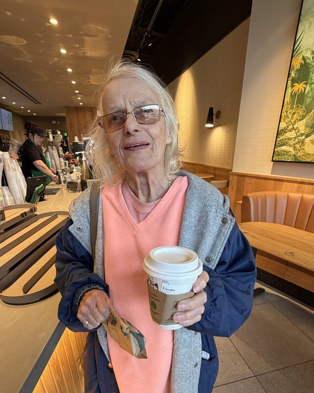 Gale Howell holding a coffee cup, wearing a coral sweater vest, grey cardigan and navy jacket.