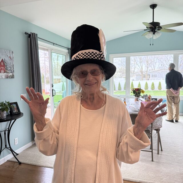 Gale Howell in a tall black top hat with a feather and checkered band, hands raised, in a pale blue room.