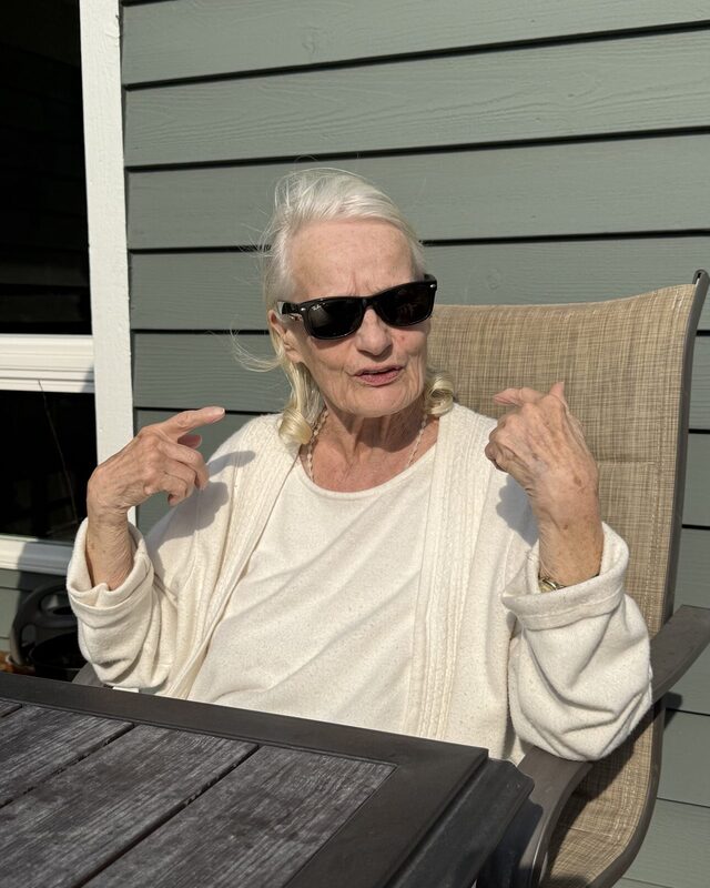 Gale Howell sitting outdoors in bright sunlight, wearing dark sunglasses and a cream sweater, gesturing with both hands against sage-green siding.